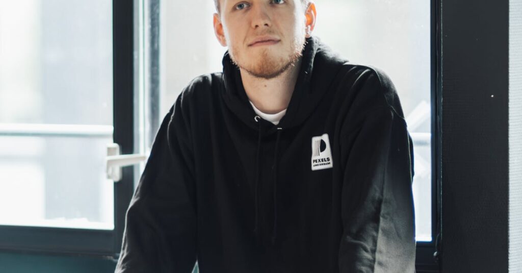 A man in a black hoodie is sitting by a window indoors, looking thoughtful and relaxed.